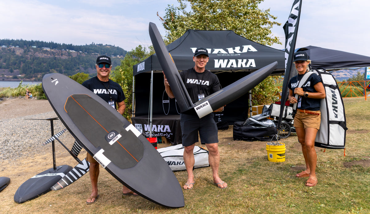 Three people holding Waka Waka surfboards near a branded tent by a body of water.